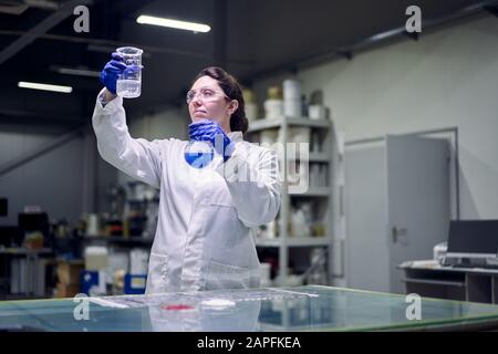 Bruna ragazza da laboratorio in occhiali e camice bianco con matraccio con liquido blu nelle sue mani conduce esperimenti su fondo defocused Foto Stock