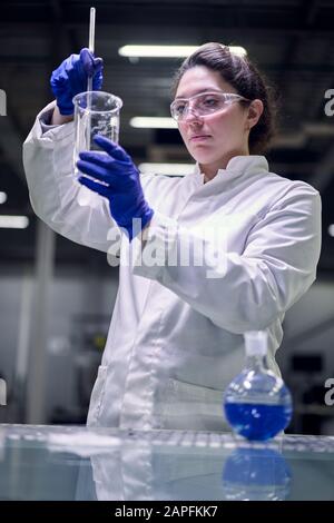 Giovane ragazza di laboratorio in occhiali e camice bianco con vetro sperimentale nelle sue mani conduce esperimenti su sfondo defocused Foto Stock