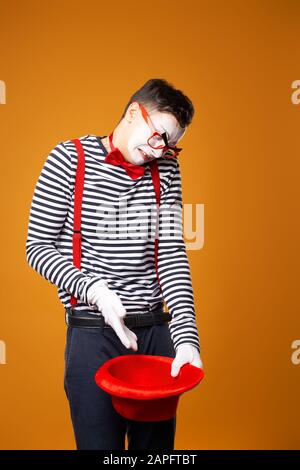 mimo triste in gilet tenendo vuoto cappello rosso isolato su sfondo arancione Foto Stock