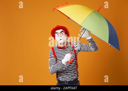 mimo triste guardando la macchina fotografica con ombrello multicolore in mano isolato su sfondo arancione vuoto in studio Foto Stock