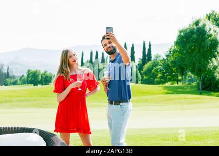 Coppia con bicchieri da vino che prendono selfie sul campo da golf Foto Stock