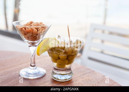 Olive verdi con limone e ghiaccio e arachidi in bicchieri su un tavolo rotondo in legno, antipasto gratuito in un bar, foto con fuoco selettivo Foto Stock