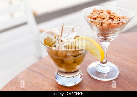 Olive verdi con limone e ghiaccio e arachidi in due bicchieri su un tavolo rotondo in legno, antipasto gratuito in un bar, fotografia di primo piano con selezione Foto Stock