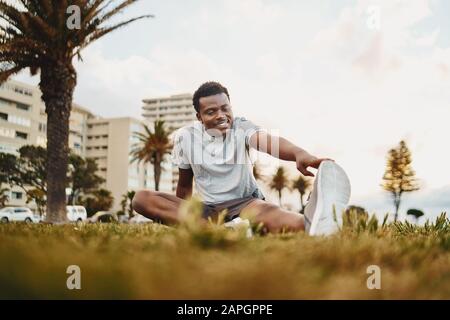 Ritratto sorridente di un giovane atleta seduto su erba verde facendo esercizio stretching al parco Foto Stock