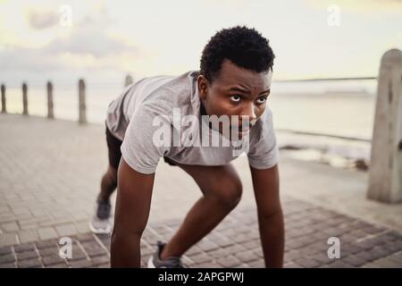 Ritratto di un giovane atleta africano americano maschio in posizione di partenza in preparazione per la corsa lungo il mare Foto Stock
