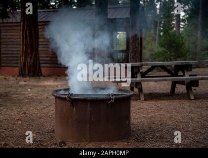 Cabina di legno standard campeggio in un campeggio California state Park, con una panca di legno e un anello di fuoco con fumo proveniente da esso Foto Stock