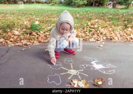 Bambino 3-5 anni, felice disegna pastelli colorati sul pavimento, creatività nel parco, sentirsi felice. Abbigliamento casual caldo con cappuccio. Giorno d'autunno. Gratis Foto Stock