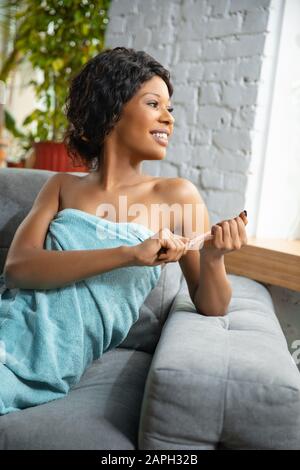 Giorno Di Bellezza. Donna afro-americana in asciugamano preparato per fare la sua routine quotidiana di cura della pelle a casa. Sedendosi sul divano, facendo manicure, sorridendo. Concetto di bellezza, cura di sé, cosmetici, giovani, sani. Foto Stock