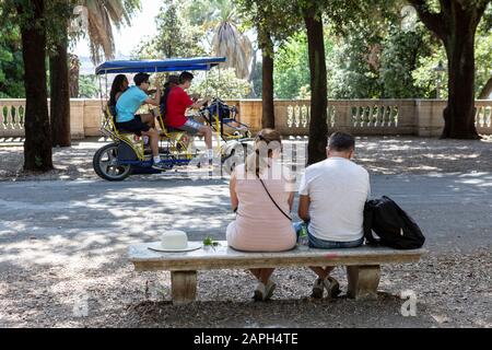 Coppia seduta su una panchina di un parco in pietra a Villa Borghese a Roma, mentre quattro persone fanno un giro in bici Surrey sullo sfondo. Foto Stock