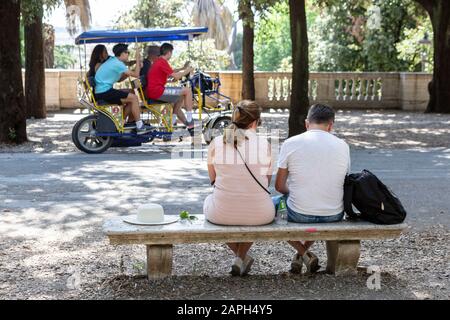 Coppia seduta su una panchina di un parco in pietra a Villa Borghese a Roma, mentre quattro persone fanno un giro in bici Surrey sullo sfondo. Foto Stock