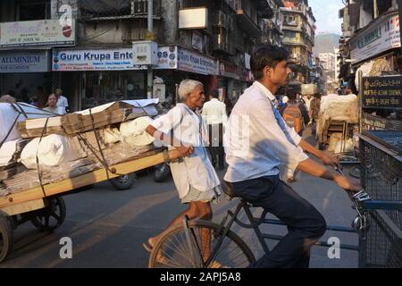 Nella trafficata Kalbadevi Road, Bhuleshwar, Mumbai, India, un portiere lotta per tirare il suo pesante carrello attraverso il traffico caotico, sorvolato da un ciclista Foto Stock