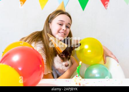 Carino piccolo cane divertente con una torta di compleanno e un cappello festa che celebra il compleanno con ragazza amante. Bella giovane donna abbraccia un cane in tappi di vacanza. Festa di compleanno del cane. Concetto di amicizia. Foto Stock