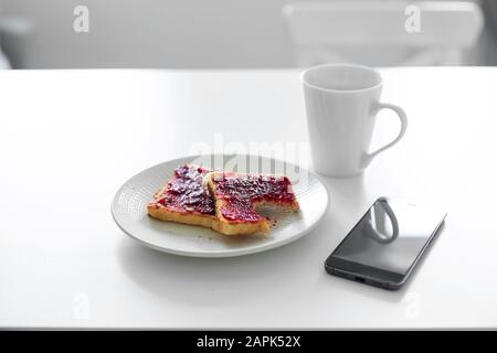 La colazione è a tavola. Una tazza di caffè e toast con marmellata di ciliegie accanto al telefono. Foto Stock