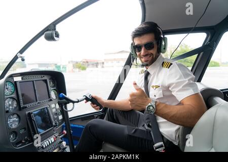 Ritratto di bel pilota commerciale in uniforme bianca capitano con cuffia seduta e pollice in su e l'avvio dei comandi nel cockpit in privato Foto Stock