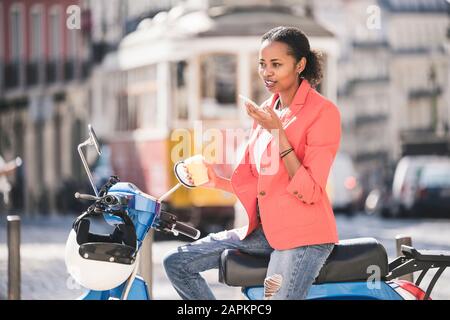 Giovane donna con scooter a motore utilizzando cellulare nella città, Lisbona, Portogallo Foto Stock