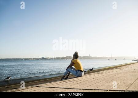 Vista posteriore della giovane donna seduta sul lungomare al tramonto, Lisbona, Portogallo Foto Stock