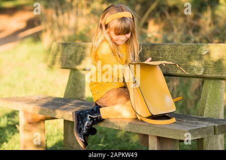 Ragazza bionda piccola vestita di giallo seduta su panca da giardino con borsa in pelle Foto Stock