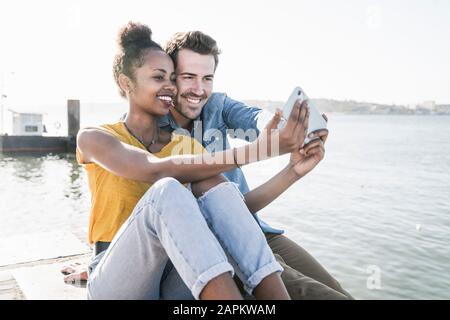 Felice giovane coppia seduto sul molo sul lungomare prendendo un selfie, Lisbona, Portogallo Foto Stock