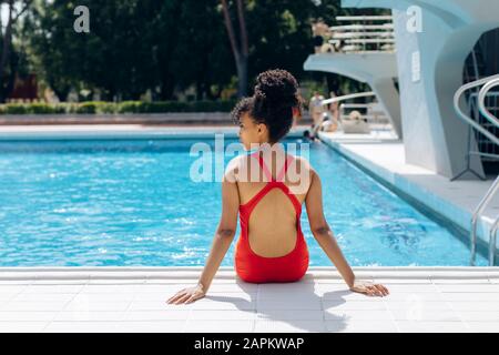 Vista posteriore della giovane donna che indossa un costume da bagno rosso seduto a bordo piscina Foto Stock