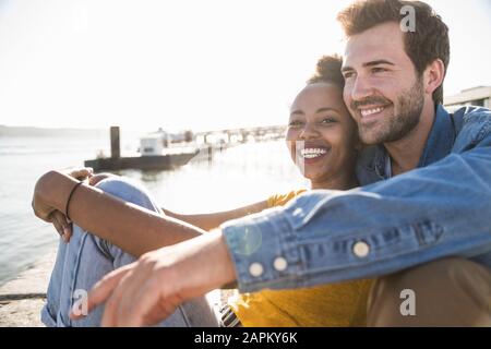 Ritratto di felice giovane coppia seduto sul molo sul lungomare, Lisbona, Portogallo Foto Stock
