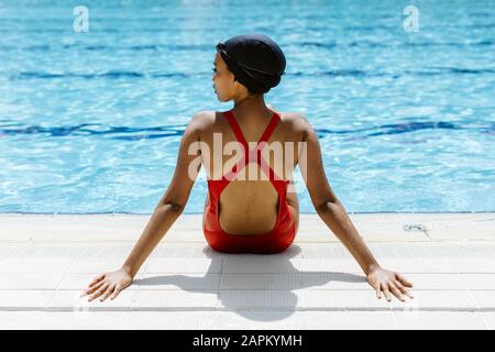 Vista posteriore di una giovane donna che indossa un costume da bagno rosso e un berretto da bagno rilassante a bordo piscina dopo il bagno Foto Stock