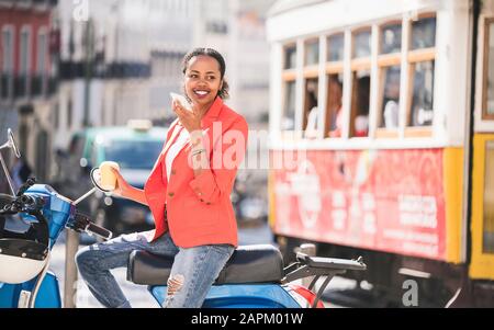 Giovane donna sorridente con scooter a motore che usa il cellulare nella città, Lisbona, Portogallo Foto Stock