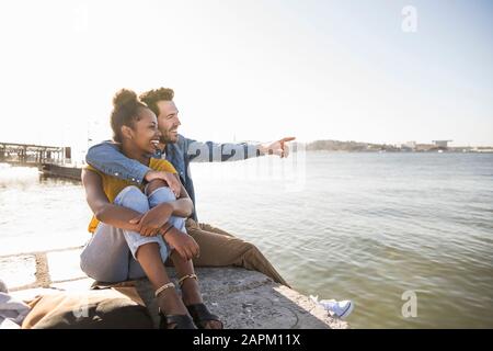Felice giovane coppia seduto sul molo in riva al mare vista, Lisbona, Portogallo Foto Stock