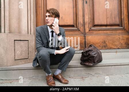 Ritratto di giovane uomo d'affari sul telefono seduto su gradini di ingresso della casa Foto Stock