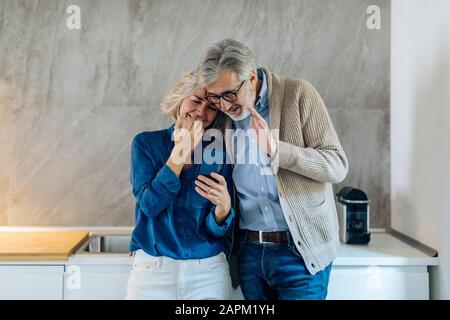 Coppia matura usando il telefono delle cellule insieme in cucina a casa Foto Stock
