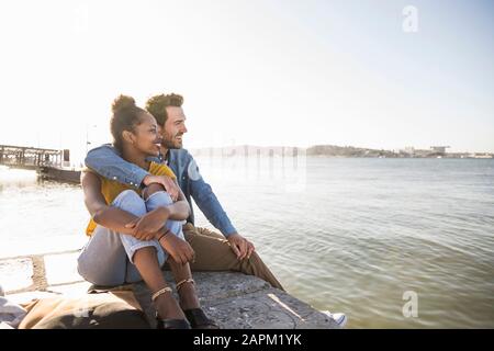 Giovane coppia seduto sul molo sul lungomare godendo della vista, Lisbona, Portogallo Foto Stock
