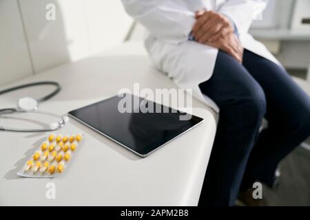 Medico seduto sul tavolo nella sua pratica medica con compressa, stetoscopio e capsule Foto Stock