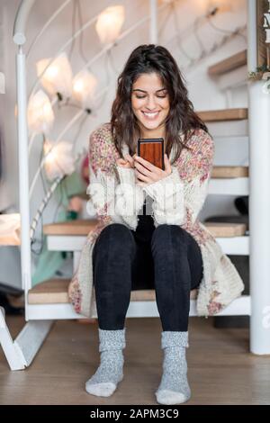 Ritratto di giovane donna felice seduto sulle scale a casa utilizzando lo smartphone Foto Stock