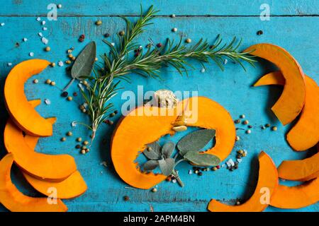 Fette di kuri squash rosso (Cucurbita maxima), rosmarino, salvia, sale e pepe adagiate sul tagliere blu Foto Stock