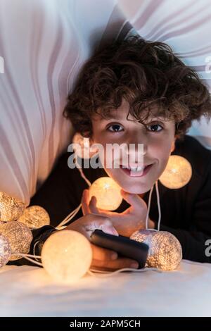 Ritratto di ragazzo sorridente con una catena di luci e telefono cellulare sotto la copertura del letto Foto Stock
