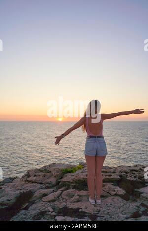 Giovane donna in piedi sulla scogliera, guardando il tramonto, vista posteriore Foto Stock