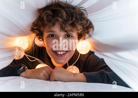 Ritratto di ragazzo felice con una catena di luci sotto il copriletto Foto Stock