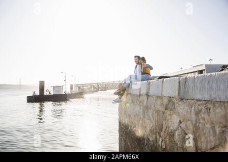 Giovane coppia seduto sul molo sul lungomare godendo della vista, Lisbona, Portogallo Foto Stock