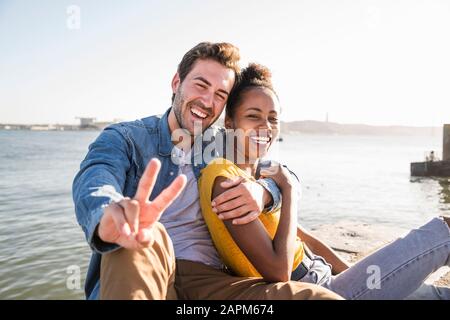 Ritratto di felice giovane coppia seduto sul lungomare gesturing, Lisbona, Portogallo Foto Stock