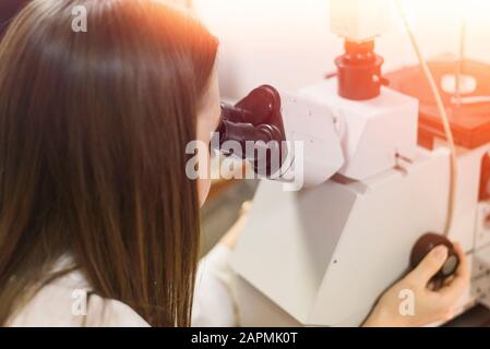 Una brunetta in un camice da laboratorio bianco guarda attraverso un microscopio in laboratorio. Un ricercatore esamina il metallo. Donna nella scienza. Foto Stock