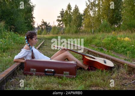 Una ragazza adolescente è seduto sui binari con una chitarra e una vecchia valigia Foto Stock