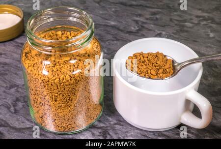 Un cucchiaio di granuli di caffè istantaneo da un vasetto di conservazione in vetro che viene posto in una tazza bianca semplice. Foto Stock