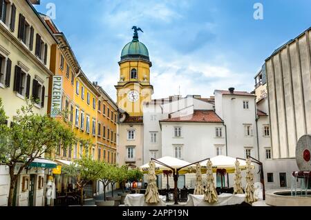 Rijeka, Croazia - 19 maggio 2019: Piazza dietro la Torre dell'Orologio della Città gialla con tavoli da caffè, ombrelloni e ulivi in una giornata di sole. Edifici gialli con Foto Stock