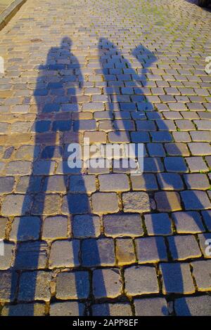 Ombre scure e allungate di uomo e donna su un vecchio pavimento di ciottoli in una giornata di sole. Le pietre sono colorate e riflettono i raggi del sole. Foto Stock