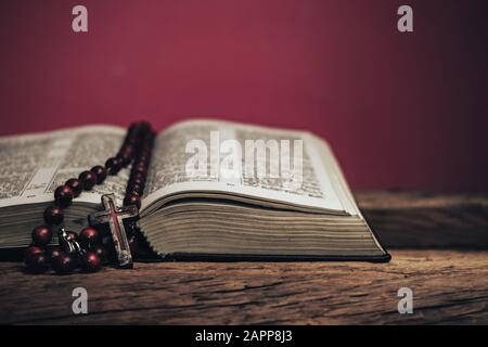 Apri la Sacra Bibbia e beads crocifisso su un vecchio tavolo di legno di quercia. Bellissimo sfondo rosso muro. Foto Stock