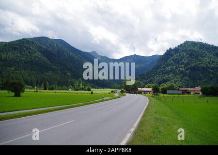 Una strada asfaltata vuota attraverso i campi verdi che conducono verso il villaggio di montagna. In lontananza, ci sono montagne verdi fortemente boscose. Foto Stock