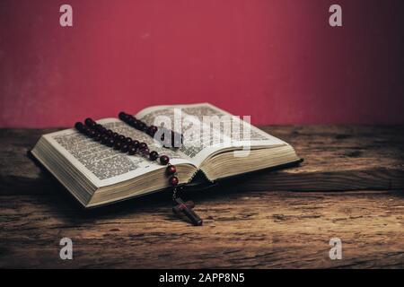 Apri la Sacra Bibbia e beads crocifisso su un vecchio tavolo di legno di quercia. Bellissimo sfondo rosso muro. Foto Stock