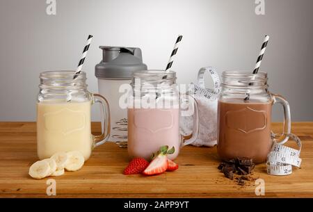 Milkshake di banana, fragola e cioccolato in bicchieri da Vasetto Mason, con cannucce di carta, ingredienti e shaker su un piano di lavoro in legno Foto Stock