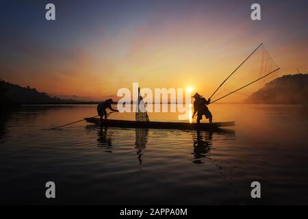 Silhouette di pescatore asiatico su barca di legno in azione gettando una rete per catturare pesci d'acqua dolce nella natura fiume al mattino presto prima dell'alba Foto Stock