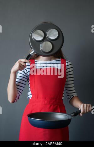 L'immagine di una giovane donna in un grembiule cuoceva il suo viso con una padella per cucinare frittelle, tiene la padella ai livelli della cintura con la sua seconda h. Foto Stock