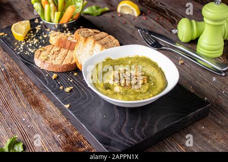 Hummus con spinaci, avocado e semi di zucca in una ciotola su una tavola di legno e bruschetta, cucina orientale e araba Foto Stock